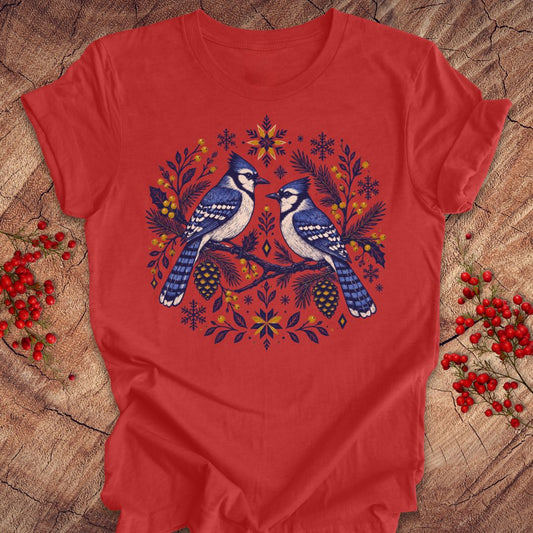 A red short-sleeve crewneck t-shirt laid flat on a rustic wood background, featuring a colorful graphic of two bluebirds surrounded by pinecones, pine branches, and snowflakes in blue, yellow, and orange.