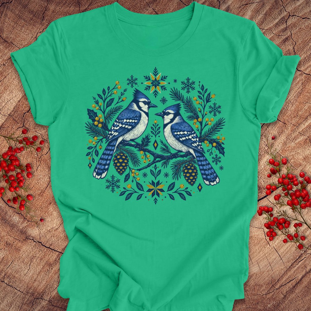 A bright green short-sleeve crewneck t-shirt featuring a detailed graphic of two blue jays surrounded by pinecones, pine needles, and snowflakes in a circular wreath design. The shirt is laid flat on a rustic wood surface decorated with red berries.