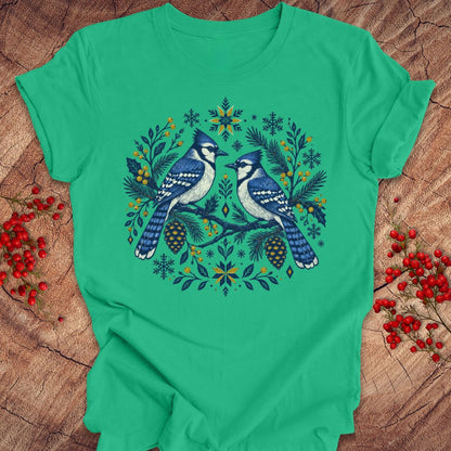 A bright green short-sleeve crewneck t-shirt featuring a detailed graphic of two blue jays surrounded by pinecones, pine needles, and snowflakes in a circular wreath design. The shirt is laid flat on a rustic wood surface decorated with red berries.