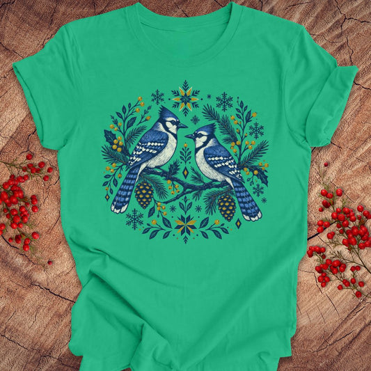 A bright green short-sleeve crewneck t-shirt featuring a detailed graphic of two blue jays surrounded by pinecones, pine needles, and snowflakes in a circular wreath design. The shirt is laid flat on a rustic wood surface decorated with red berries.