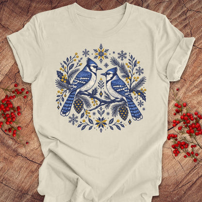 An off-white crewneck t-shirt featuring a detailed graphic of two blue jays surrounded by pine branches, pinecones, and snowflakes in blue, yellow, and white. The t-shirt is laid flat on a rustic wood background decorated with red berries.