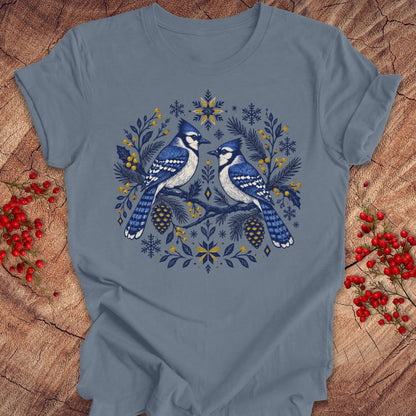 A slate blue crewneck t-shirt featuring a detailed graphic of two blue jays surrounded by pinecones, pine branches, and snowflakes in shades of blue, navy, and yellow. The t-shirt is displayed flat on a rustic wood background decorated with red berries.
