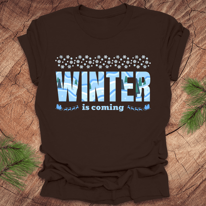 Brown t-shirt with 'Winter is coming' text on a wooden surface with pine branches.