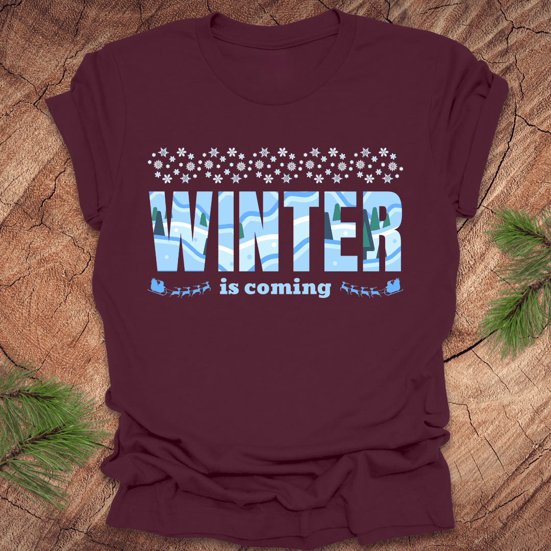Maroon t-shirt with 'Winter is coming' graphic on a wooden surface with pine branches.