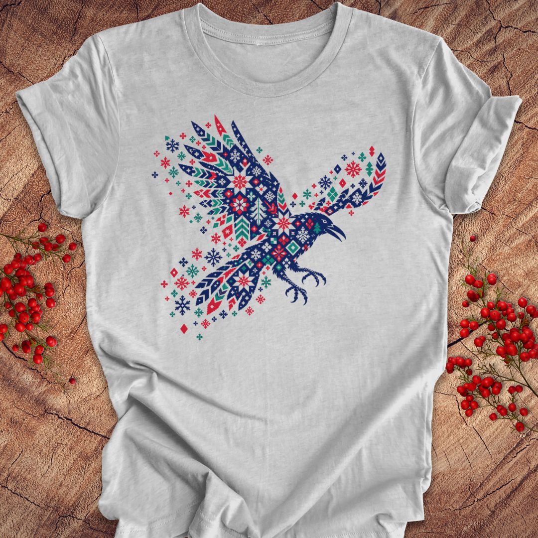 A white short-sleeve crewneck t-shirt featuring a colorful graphic of a flying raven filled with intricate red, blue, and green snowflake and geometric patterns. The shirt is displayed flat on a rustic wooden surface decorated with small red berries.