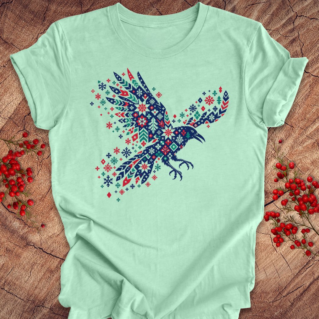 A mint green short-sleeve t-shirt featuring a colorful raven graphic filled with a Nordic-style geometric pattern in navy, red, and white. The shirt is laid flat on a rustic wood background decorated with small red berries.