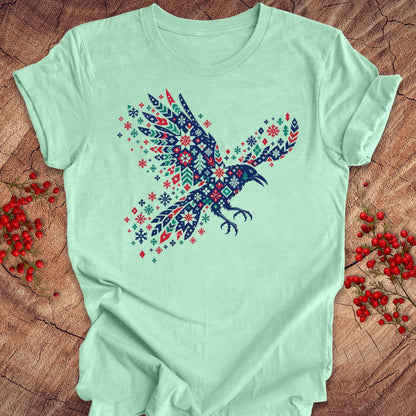 A mint green short-sleeve t-shirt featuring a colorful raven graphic filled with a Nordic-style geometric pattern in navy, red, and white. The shirt is laid flat on a rustic wood background decorated with small red berries.