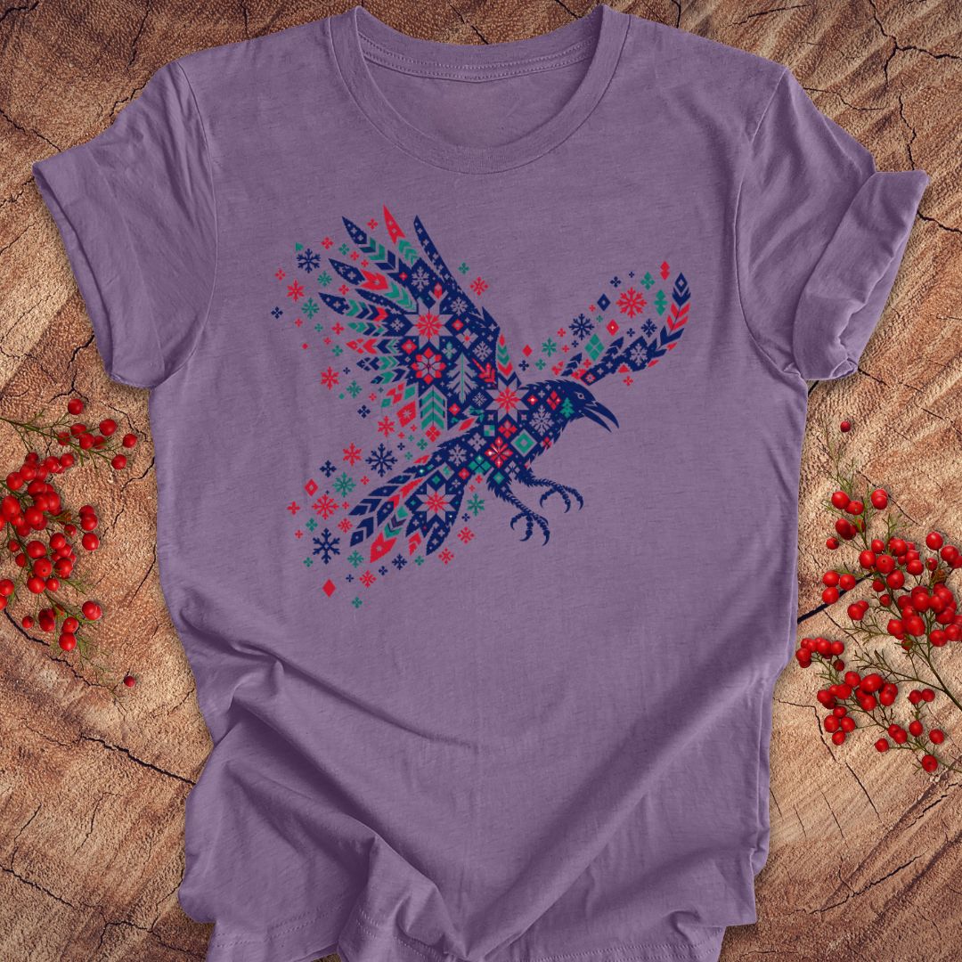 A lavender purple short-sleeve t-shirt featuring a detailed graphic of a flying raven filled with intricate snowflake and geometric patterns in navy blue, red, and teal. The t-shirt is laid flat on a rustic wood surface, flanked by small sprigs of red berries.