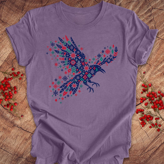 A lavender purple short-sleeve t-shirt featuring a detailed graphic of a flying raven filled with intricate snowflake and geometric patterns in navy blue, red, and teal. The t-shirt is laid flat on a rustic wood surface, flanked by small sprigs of red berries.
