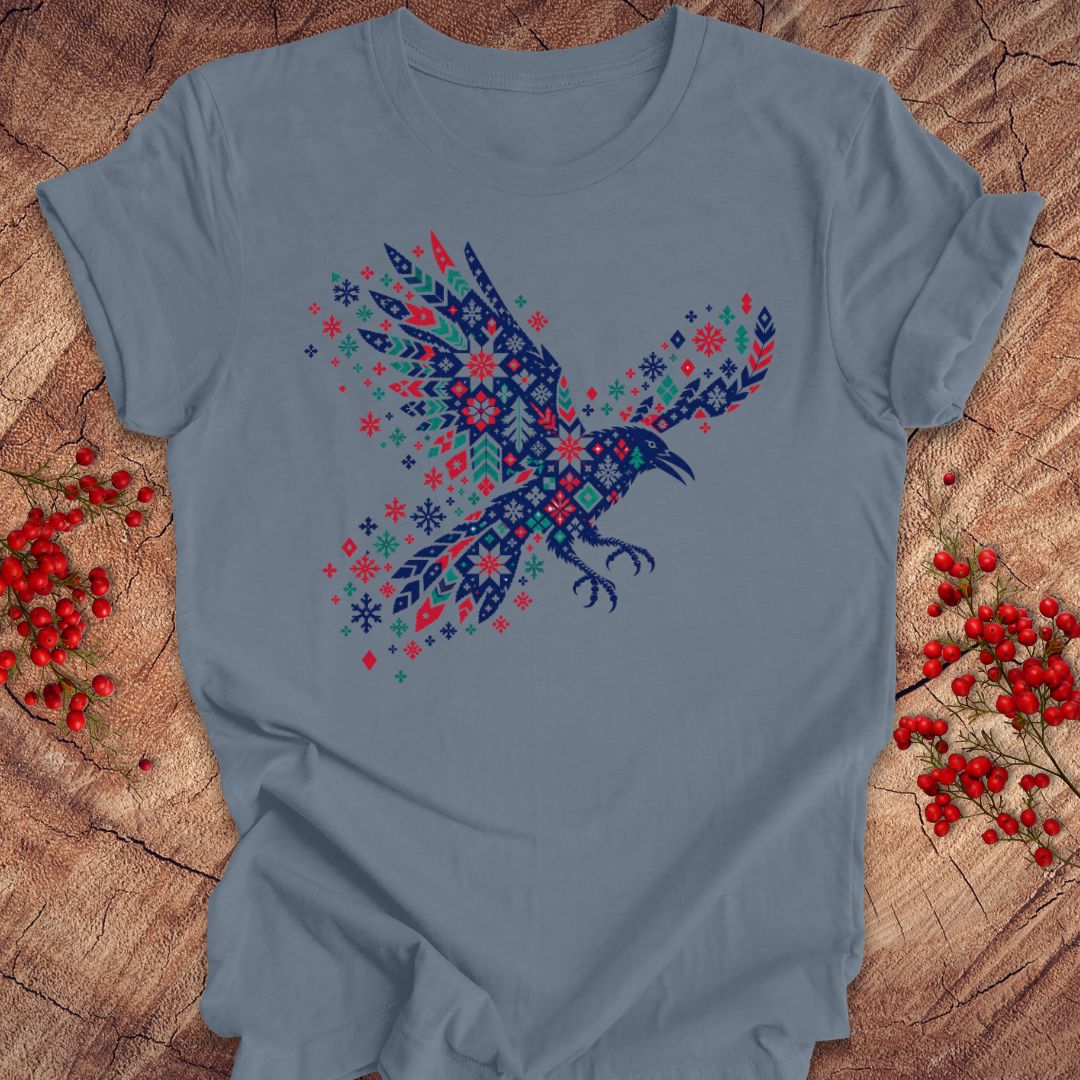 A light blue crewneck t-shirt featuring a colorful graphic of a flying raven filled with intricate geometric patterns in navy, red, and teal. The t-shirt is displayed flat on a rustic wood background decorated with small red berries.
