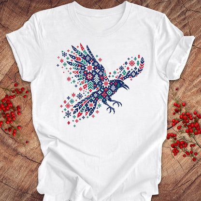 A white short-sleeve crewneck t-shirt laid flat on a rustic wood background, decorated with red and blue patterned berries on both sides. The t-shirt features a large graphic of a raven filled with intricate red, blue, and white geometric patterns.
