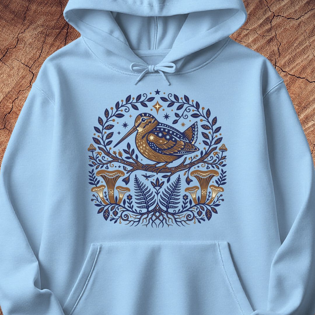 A light blue pullover hoodie laid flat on a textured brown wood background, featuring a central circular graphic of a brown bird perched on a branch surrounded by blue leaves, ferns, and orange-brown mushrooms.