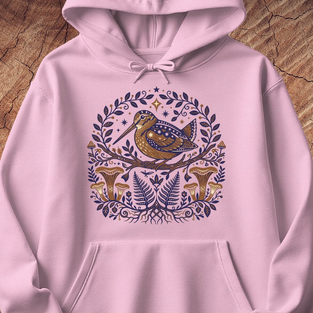 A light pink pullover hoodie laid flat on a rustic wood background, featuring a central graphic of a brown bird perched on a branch surrounded by blue foliage and gold mushrooms.