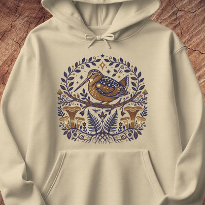 An off-white hooded sweatshirt featuring a detailed graphic print of a brown bird with blue patterned wings, surrounded by a circular wreath of blue leaves and ferns, set against a rustic wood background.
