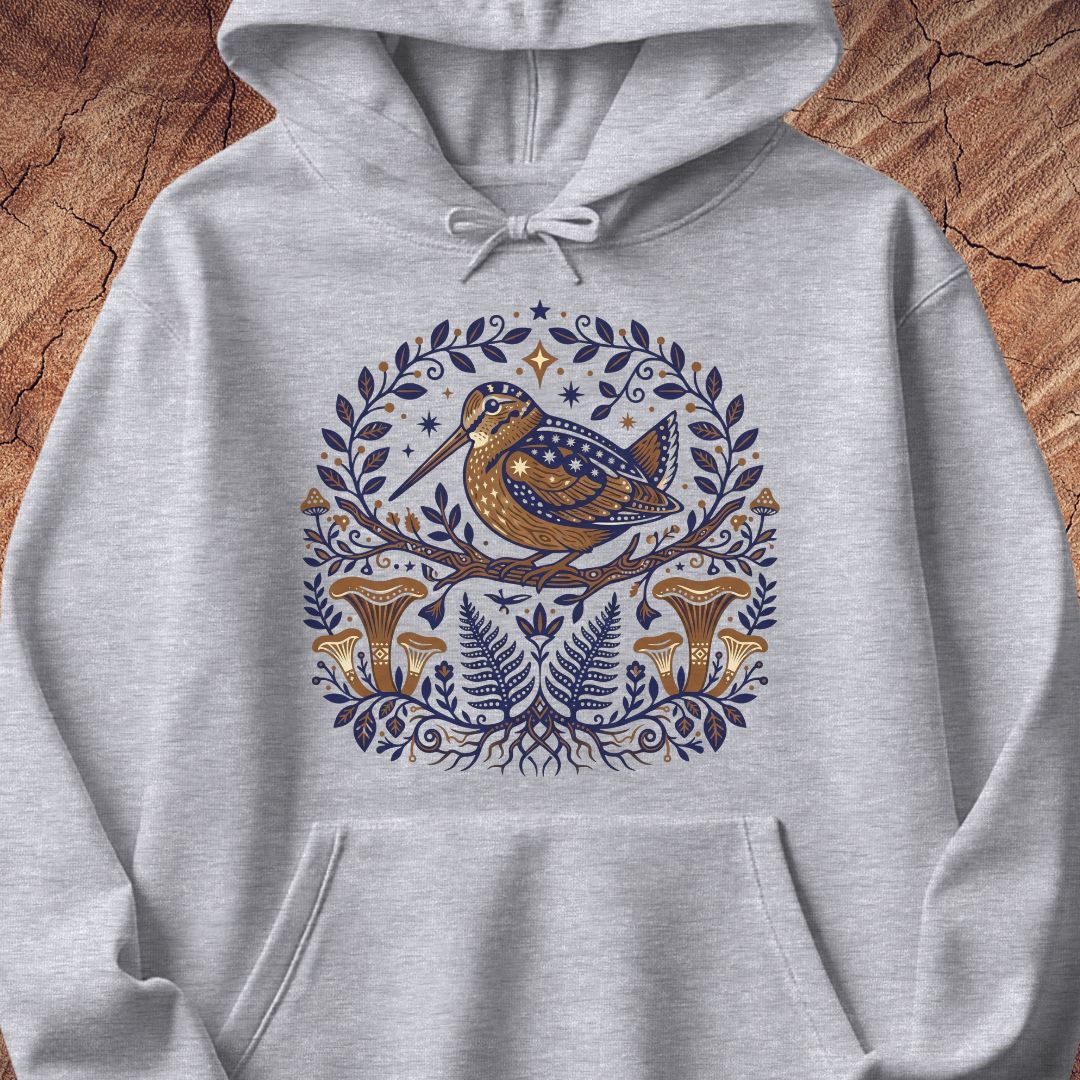 A light heather gray pullover hoodie featuring a detailed graphic print of a brown and blue bird perched on a branch, surrounded by a circular wreath of blue leaves, ferns, and gold-brown mushrooms, displayed flat against a rustic wood background.