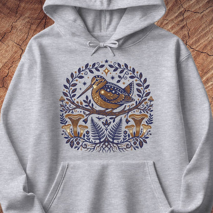 A light heather gray pullover hoodie featuring a detailed graphic print of a brown and blue bird perched on a branch, surrounded by a circular wreath of blue leaves, ferns, and gold-brown mushrooms, displayed flat against a rustic wood background.
