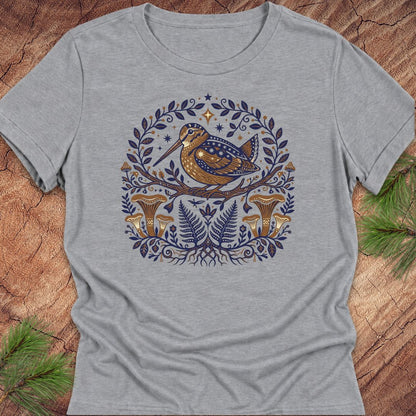 A heather gray short-sleeve t-shirt featuring a detailed graphic of a brown bird with blue spots, surrounded by a circular wreath of blue leaves, ferns, and mushrooms, laid flat on a rustic wood background with pine needles.