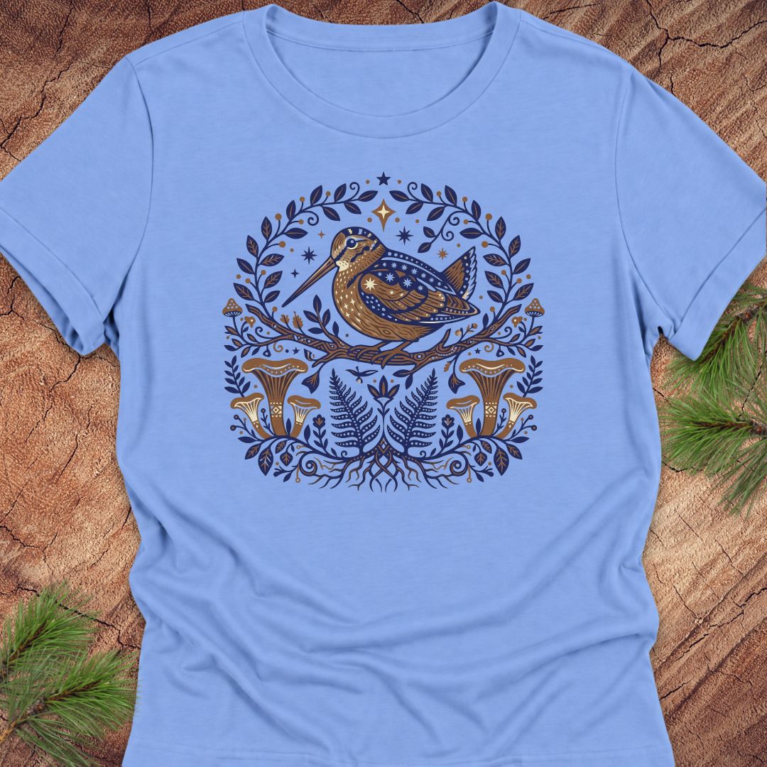 A light blue short-sleeve crewneck t-shirt featuring a detailed graphic print of a brown bird with blue spots, surrounded by a circular wreath of green leaves, pinecones, and mushrooms, laid flat on a rustic wood background with pine needles.