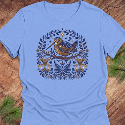 A light blue short-sleeve crewneck t-shirt featuring a detailed graphic print of a brown bird with blue spots, surrounded by a circular wreath of green leaves, pinecones, and mushrooms, laid flat on a rustic wood background with pine needles.