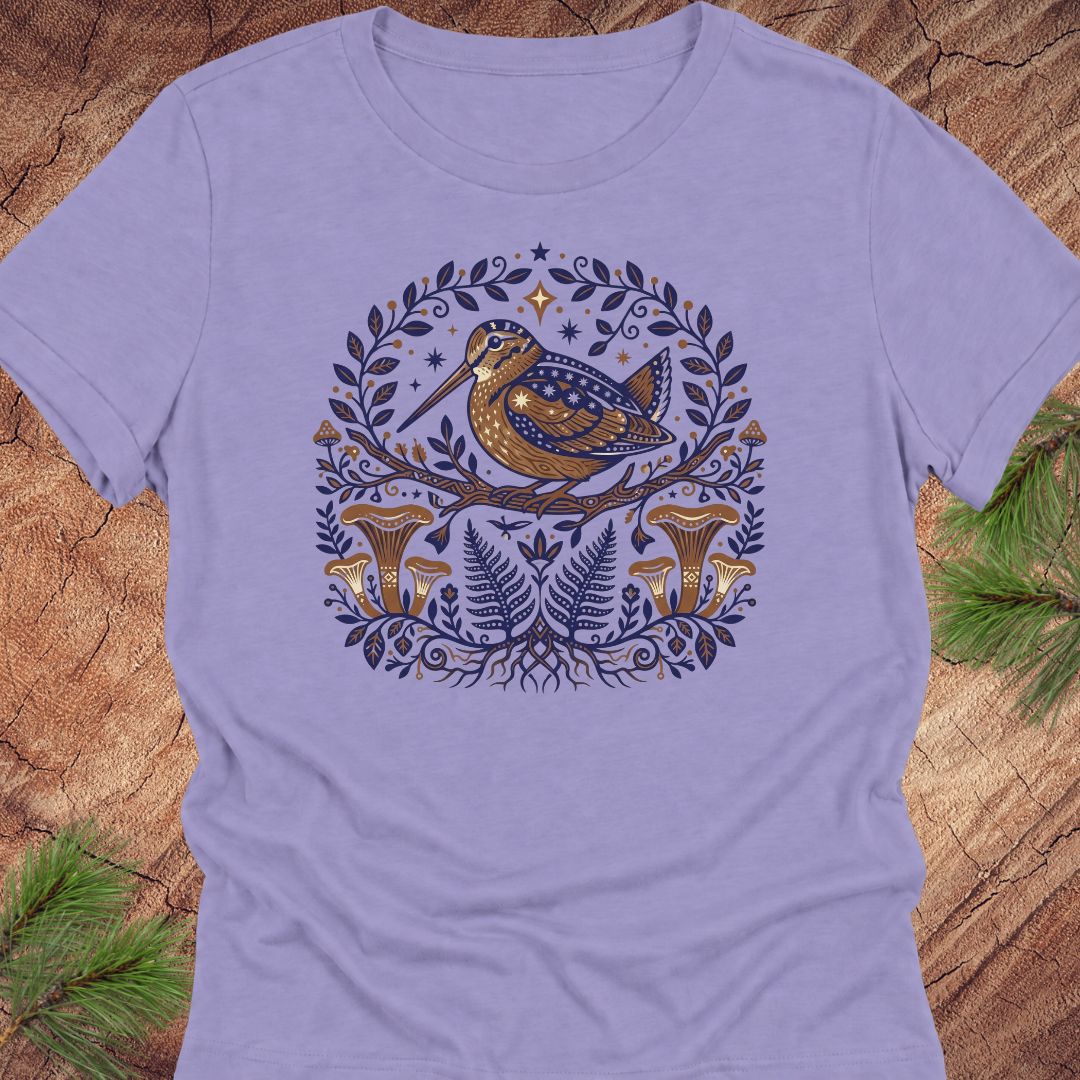 A light lavender crewneck t-shirt featuring a detailed fantasy graphic of a bird perched on a branch surrounded by foliage, mushrooms, and stars in navy blue and gold, laid flat on a rustic wood background with pine needles.