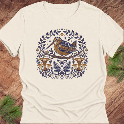 An off-white short-sleeve crewneck t-shirt featuring a detailed folk-art style graphic of a brown bird, ferns, and mushrooms in a circular frame, laid flat on a rustic wood background with pine branches.