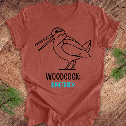 A terracotta-colored short-sleeve t-shirt with a black line art graphic of a woodcock and the text 'WOODCOCK: SHOREBIRD?' printed on the front, displayed flat on a rustic wood background with pine needles.