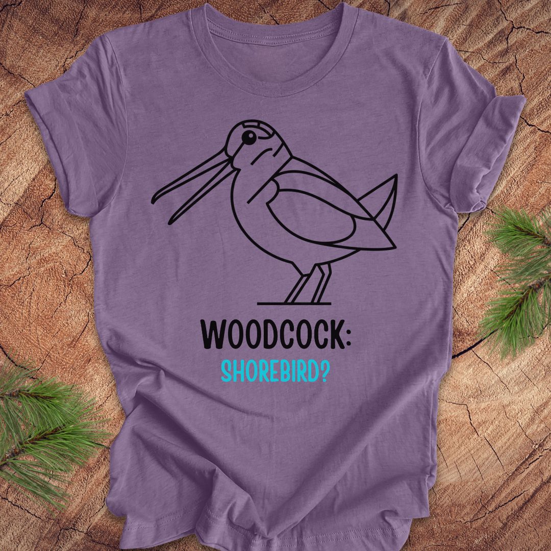 A lavender purple short-sleeve t-shirt with a black line art graphic of a woodcock and the text 'WOODCOCK: SHOREBIRD?' in black and turquoise, laid flat on a rustic wood background with pine needles in the corners.