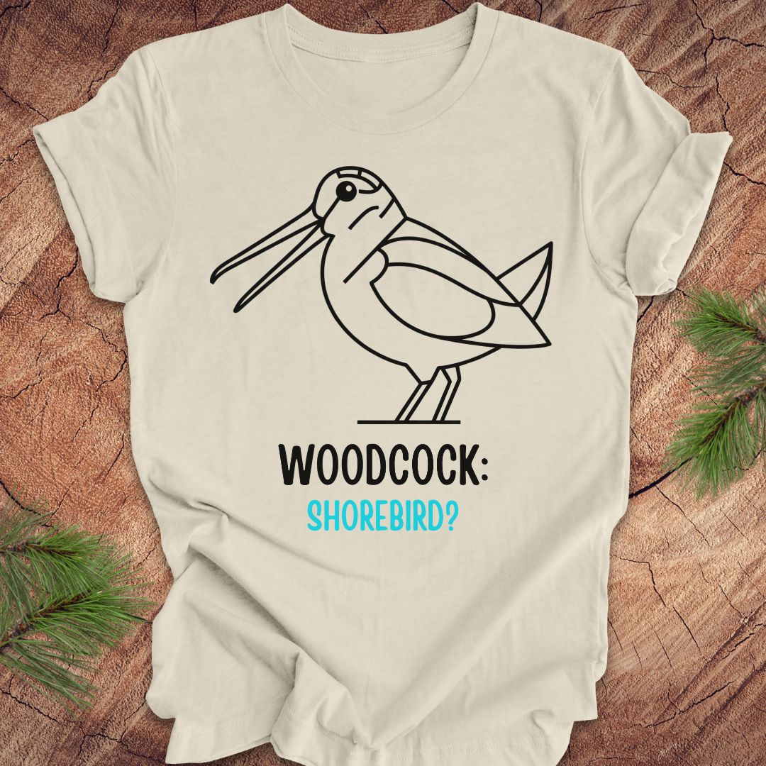 A cream-colored short-sleeve t-shirt with a black line art graphic of a woodcock and the text 'WOODCOCK: SHOREBIRD?' in black and light blue, displayed flat on a rustic wood background with pine needles.