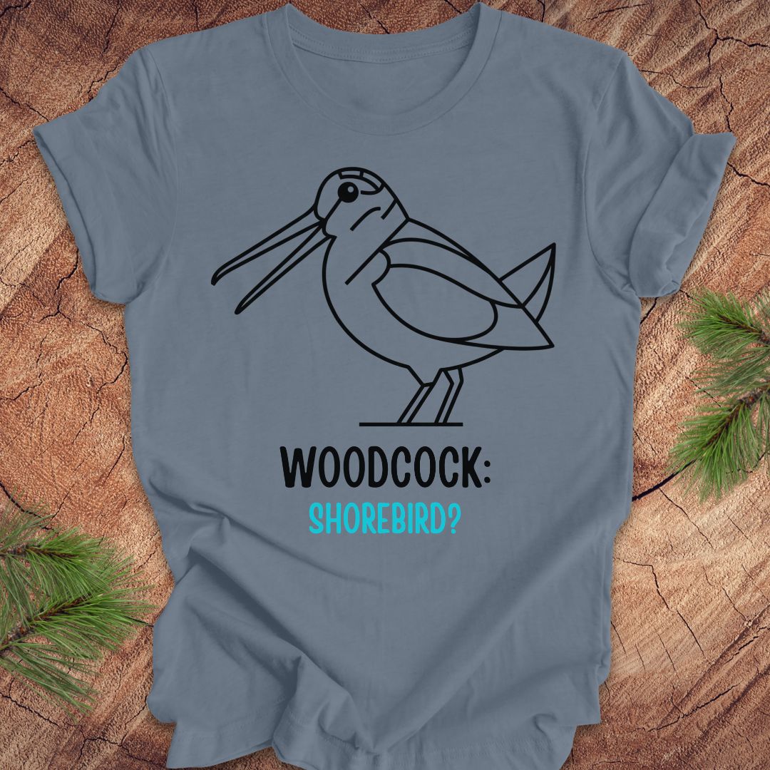 A slate blue short-sleeve t-shirt with a black line art graphic of a woodcock and the text 'WOODCOCK: SHOREBIRD?' in black and turquoise, displayed flat on a rustic wood background with pine needles.