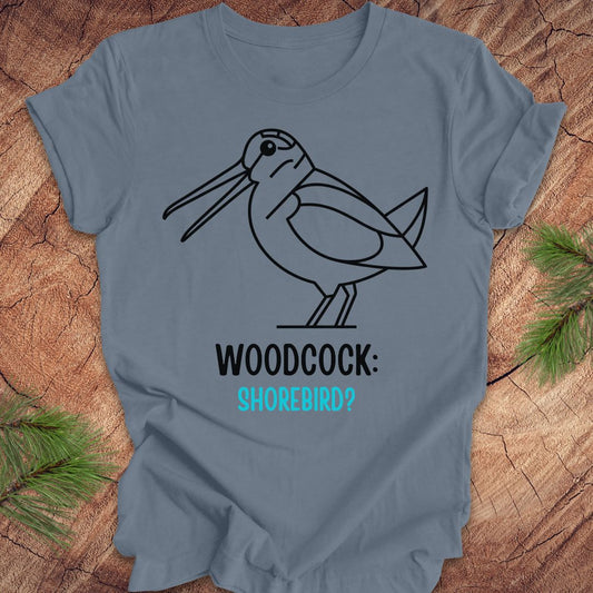 A slate blue short-sleeve t-shirt with a black line art graphic of a woodcock and the text 'WOODCOCK: SHOREBIRD?' in black and turquoise, displayed flat on a rustic wood background with pine needles.