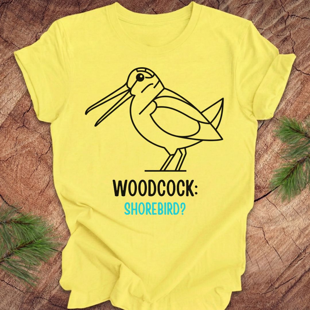 A bright yellow short-sleeve t-shirt laid flat on a rustic wood surface with pine branches at the corners. The shirt features a black line art graphic of a woodcock with the text 'WOODCOCK:' in black and 'SHOREBIRD?' in light blue.