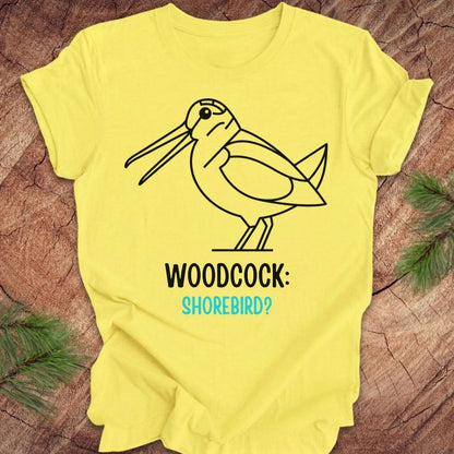 A bright yellow short-sleeve t-shirt laid flat on a rustic wood surface with pine branches at the corners. The shirt features a black line art graphic of a woodcock with the text 'WOODCOCK:' in black and 'SHOREBIRD?' in light blue.