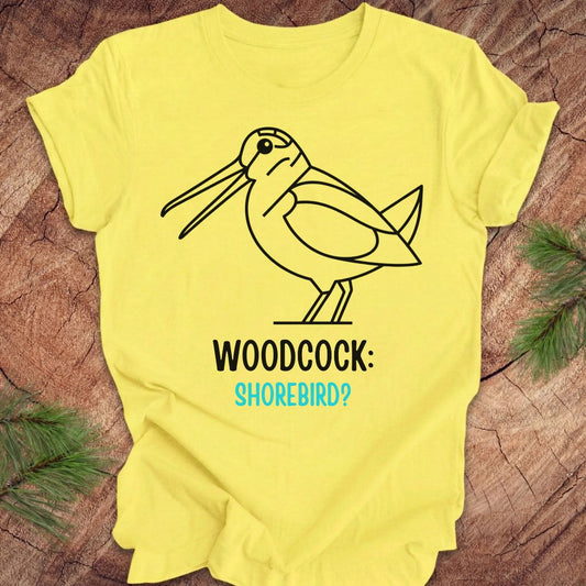 A bright yellow short-sleeve t-shirt laid flat on a rustic wood surface with pine branches at the corners. The shirt features a black line art graphic of a woodcock with the text 'WOODCOCK:' in black and 'SHOREBIRD?' in light blue.