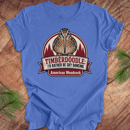 A blue short-sleeve t-shirt laid flat on a rustic wood surface with pine branches in the corners. The shirt features a large graphic of an American woodcock bird with the text 'TIMBERDOOLE: I'D RATHER BE SKY DANCING' and 'American Woodcock' on a banner.