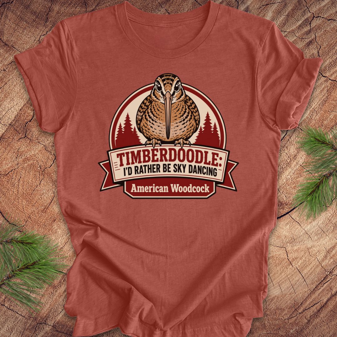 A rust-colored short-sleeve t-shirt featuring a graphic of an American Woodcock bird with the text 'TIMBERDOOLE: I'D RATHER BE SKY DANCING' and 'American Woodcock' on a banner, laid flat on a rustic wood background with pine branches.