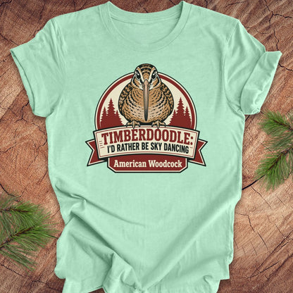 A mint green short-sleeve t-shirt with a vintage-style graphic of an American Woodcock bird and the text 'TIMBERDOOLE: I'D RATHER BE SKY DANCING' and 'American Woodcock', laid flat on a rustic wood background with pine branches.