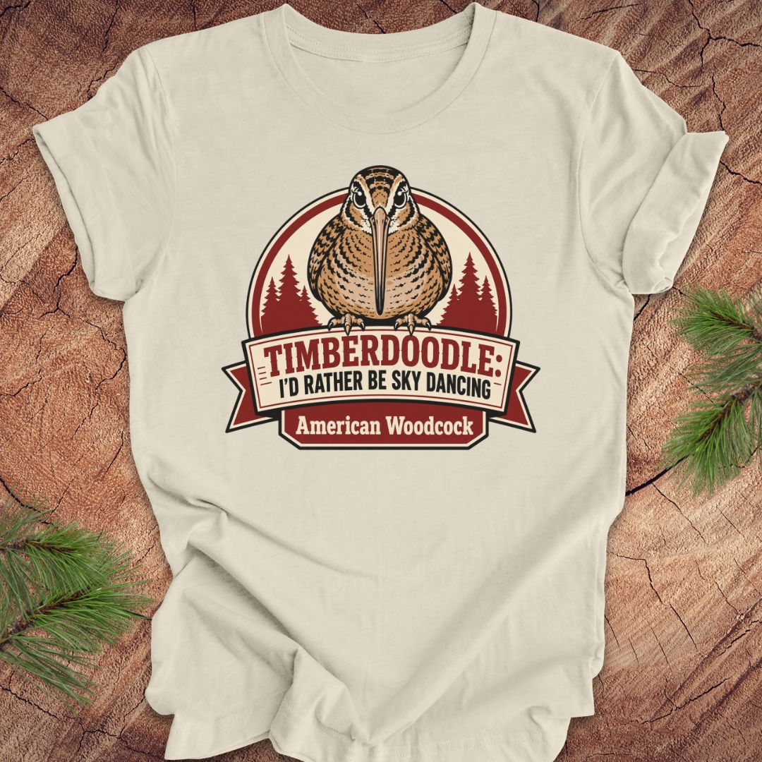 A cream-colored short-sleeve t-shirt with a graphic of an American Woodcock bird and the text 'TIMBERDOOLE: I'D RATHER BE SKY DANCING' and 'American Woodcock' on a rustic wood background with pine branches.