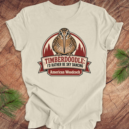 A cream-colored short-sleeve t-shirt with a graphic of an American Woodcock bird and the text 'TIMBERDOOLE: I'D RATHER BE SKY DANCING' and 'American Woodcock' on a rustic wood background with pine branches.