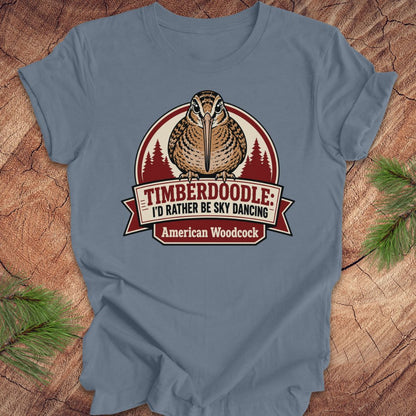 A slate blue crewneck t-shirt featuring a graphic of an American Woodcock bird with pine trees, displayed on a rustic wood background with pine needles.