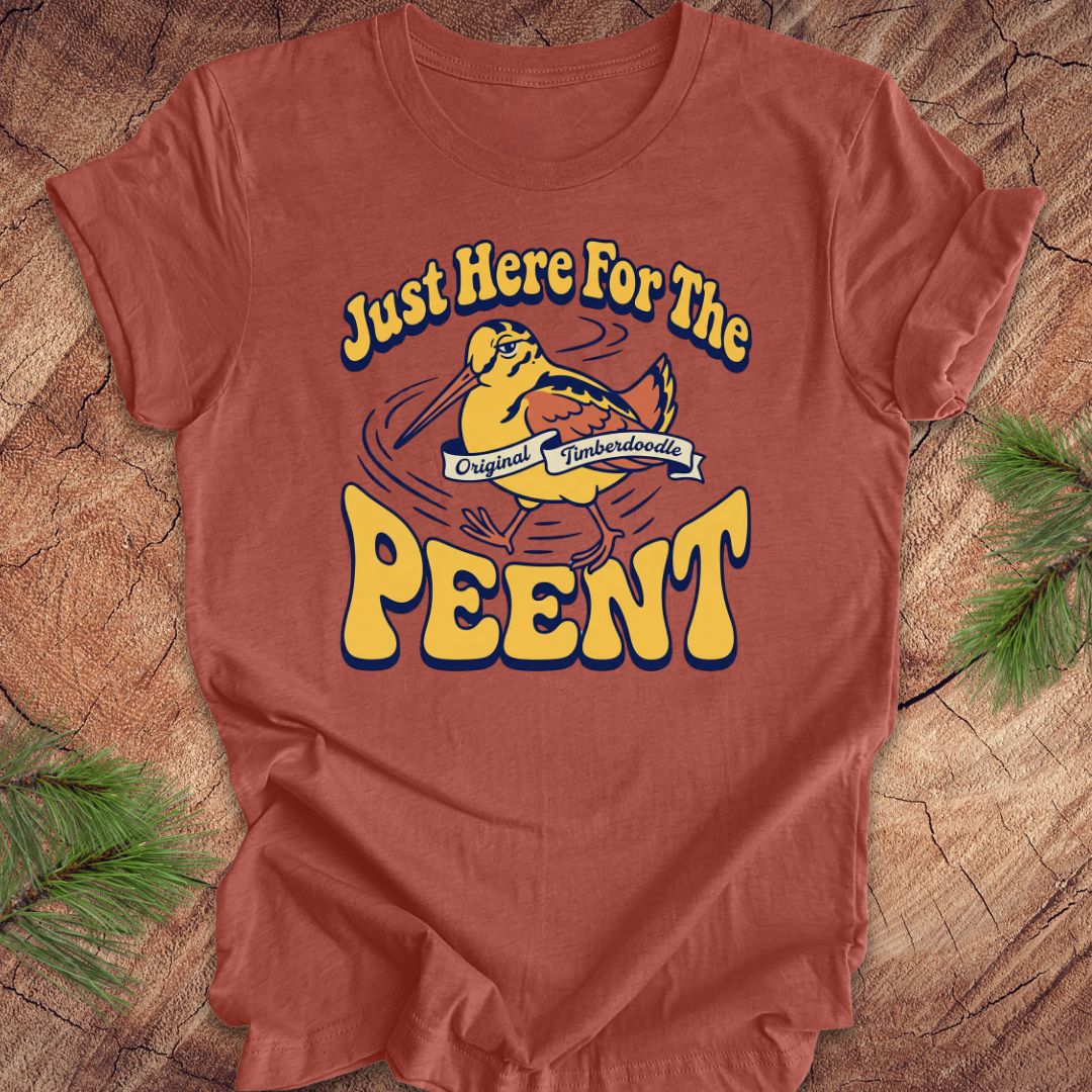 Clay colored t-shirt with 'Just Here For The Peent' woodcock graphic on a wooden surface