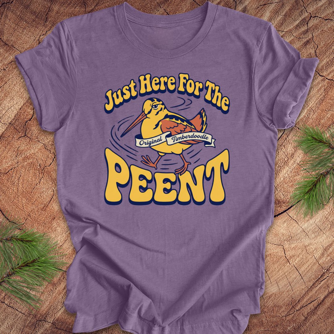 Purple t-shirt with 'Just Here For The Peent' woodcock graphic on a wooden surface