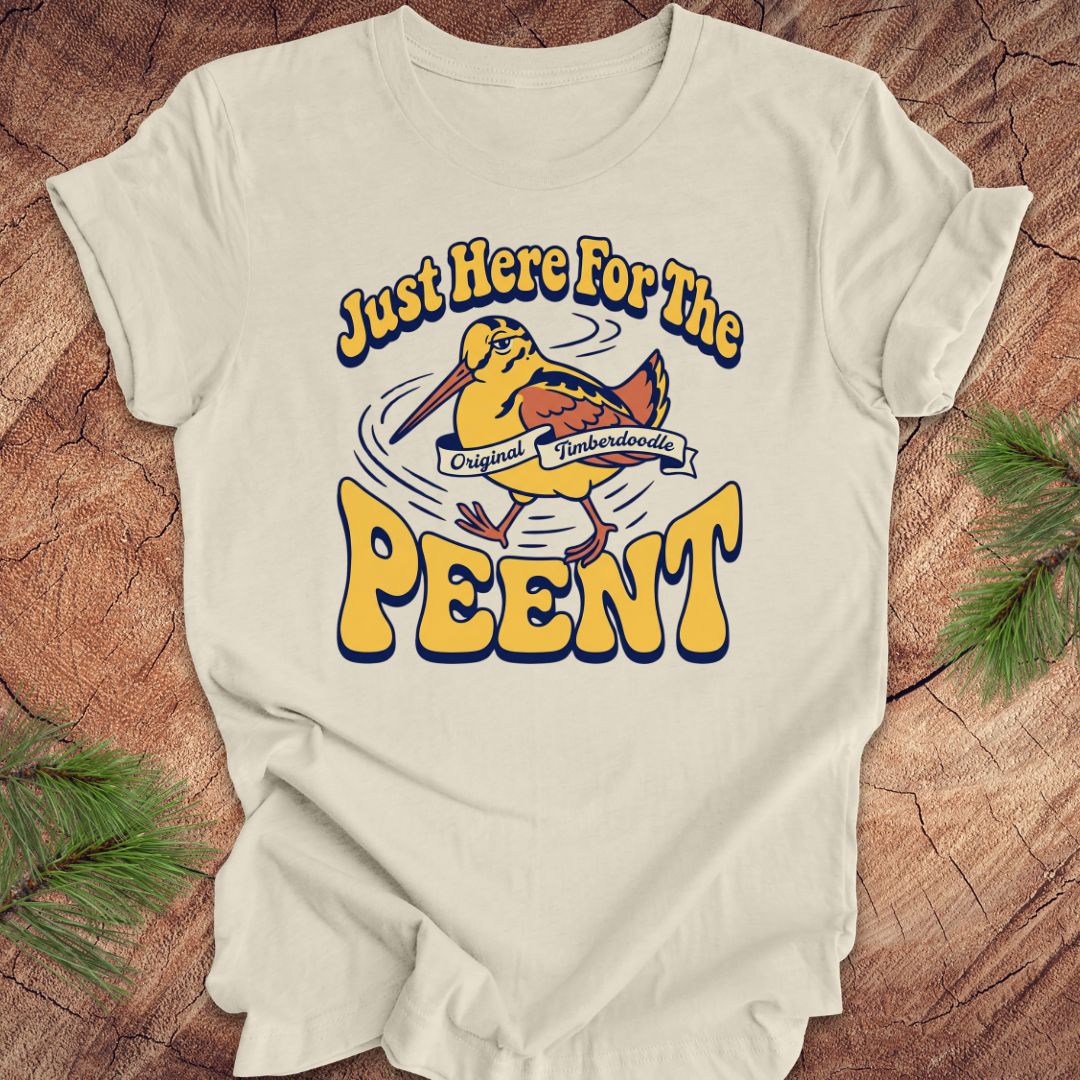 Beige t-shirt with 'Just Here For The Peent' woodcock graphic on a wooden surface