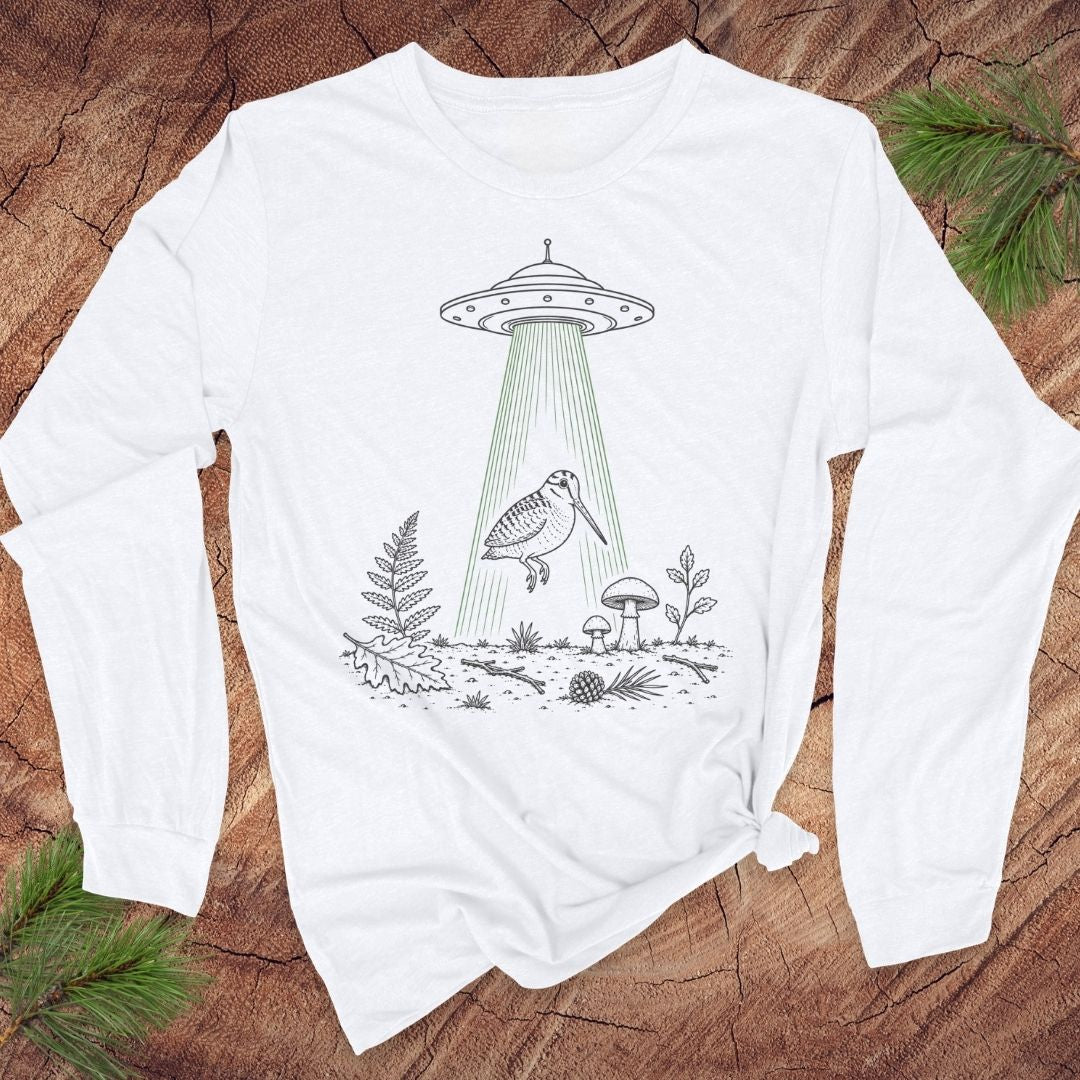 A white long-sleeve t-shirt with a black line art graphic of a UFO and a bird, styled flat on a rustic wood background with pine branches.