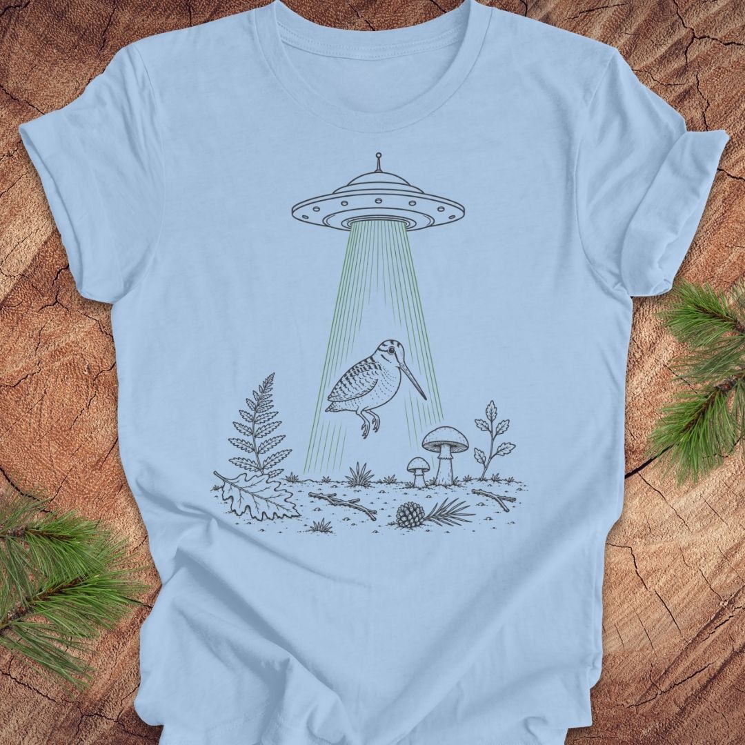 A light blue crewneck t-shirt laid flat on a rustic wood background, featuring a black line art graphic of a bird under a UFO with green beam rays.