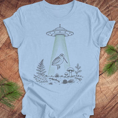 A light blue crewneck t-shirt laid flat on a rustic wood background, featuring a black line art graphic of a bird under a UFO with green beam rays.