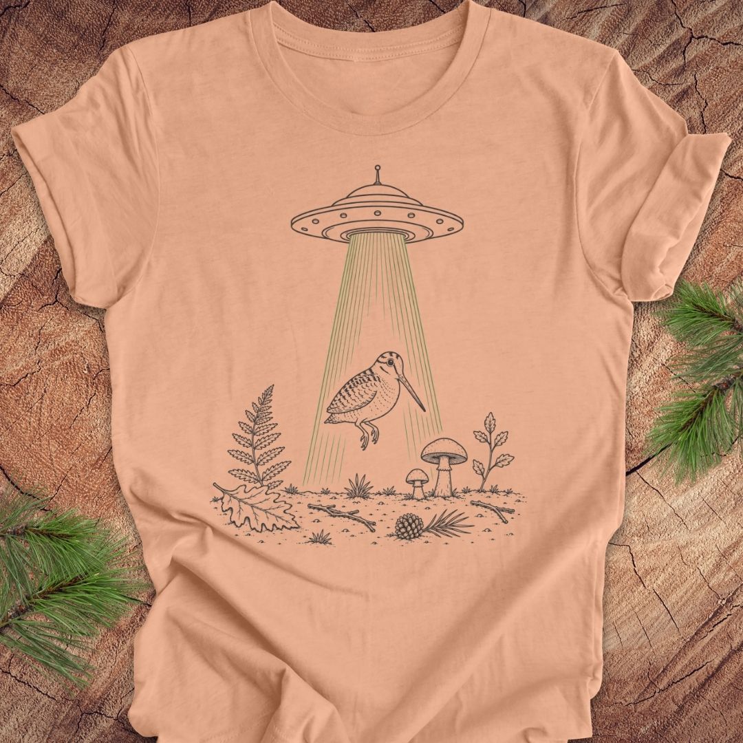 A peach-colored short-sleeve t-shirt with a graphic of a bird under a UFO beam of light, laid flat on a rustic wood background with pine branches.