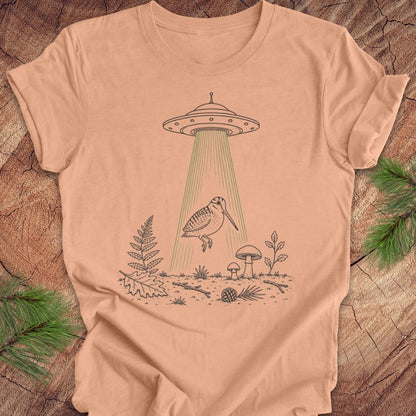 A peach-colored short-sleeve t-shirt with a graphic of a bird under a UFO beam of light, laid flat on a rustic wood background with pine branches.
