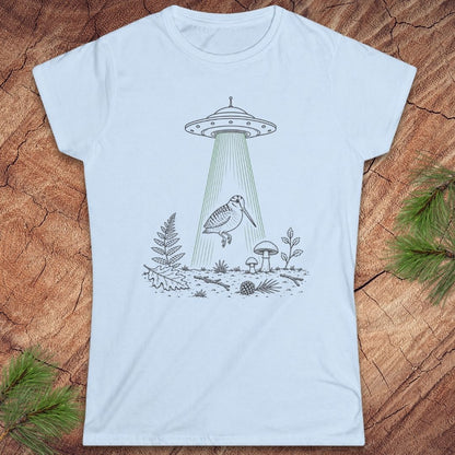 A light blue short-sleeve t-shirt with a black and green graphic of a UFO beam shining down onto a bird, ferns, and mushrooms, laid flat on a rustic wood background with pine needles in the corners.