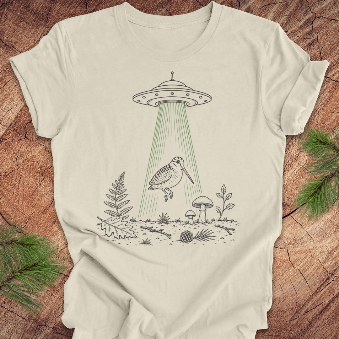 An off-white t-shirt with a black line art graphic of a UFO releasing a bird over a forest, displayed flat on a rustic wood background with pine branches.