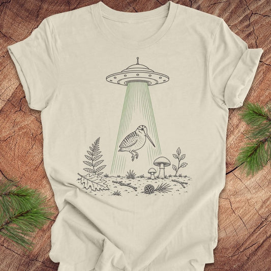 An off-white t-shirt with a black line art graphic of a UFO releasing a bird over a forest, displayed flat on a rustic wood background with pine branches.
